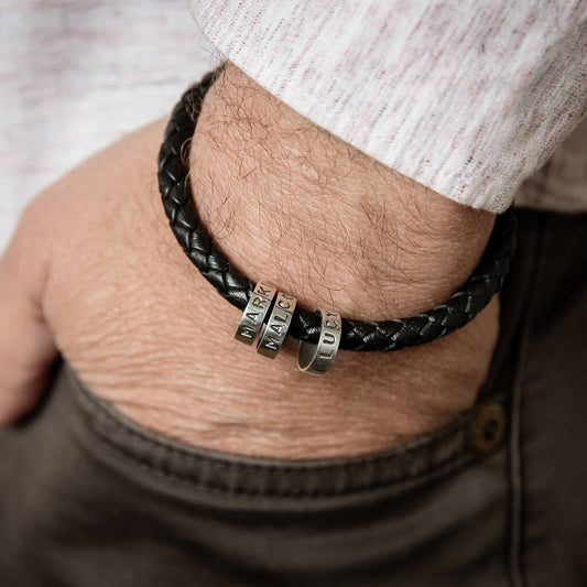 Personalised men's black braided leather bracelet with three polished sterling silver hoops hand-punched with individual names, worn on the wrist.