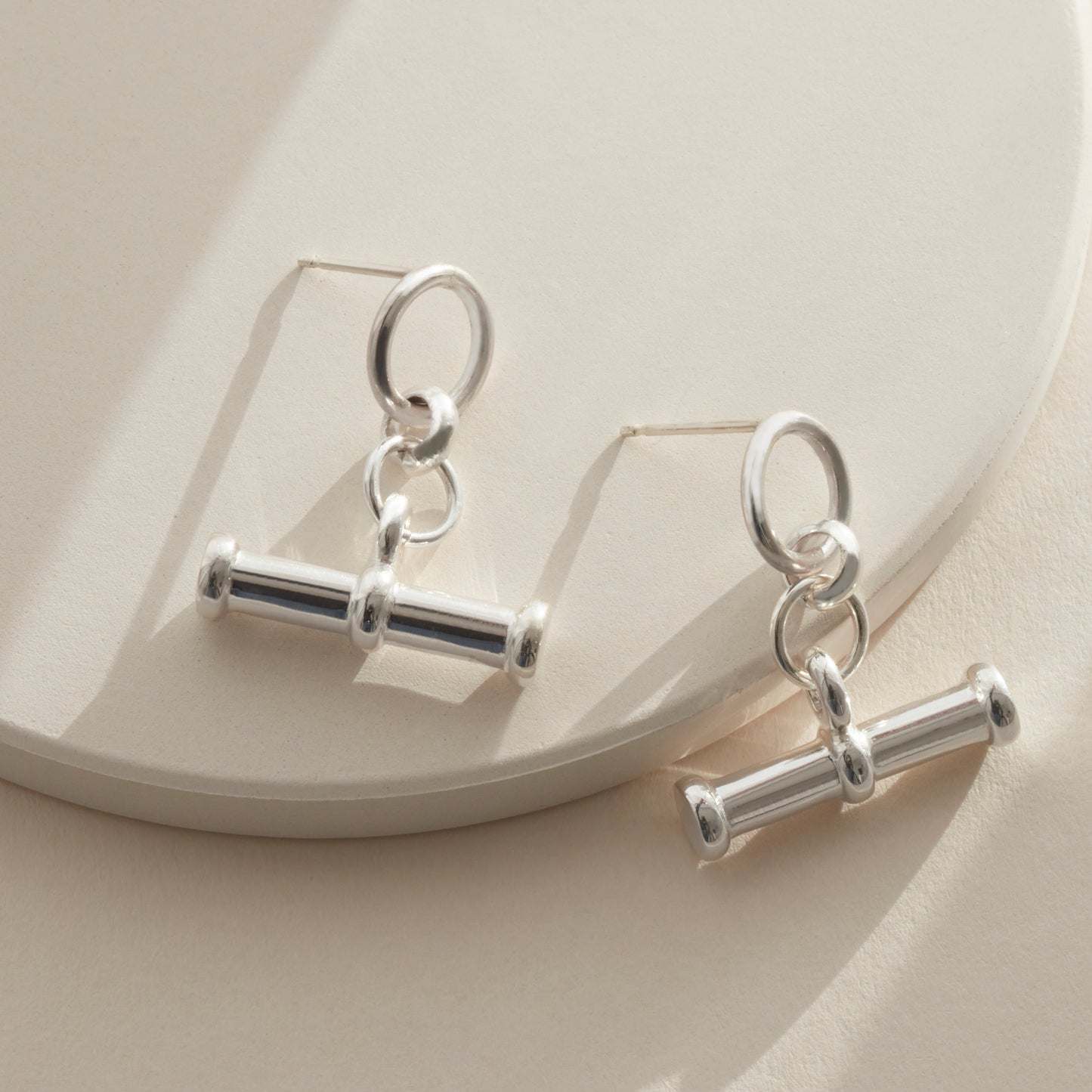 Close-up of sterling silver T bar drop earrings with a round stud and bar charm, photographed against a soft cream background.