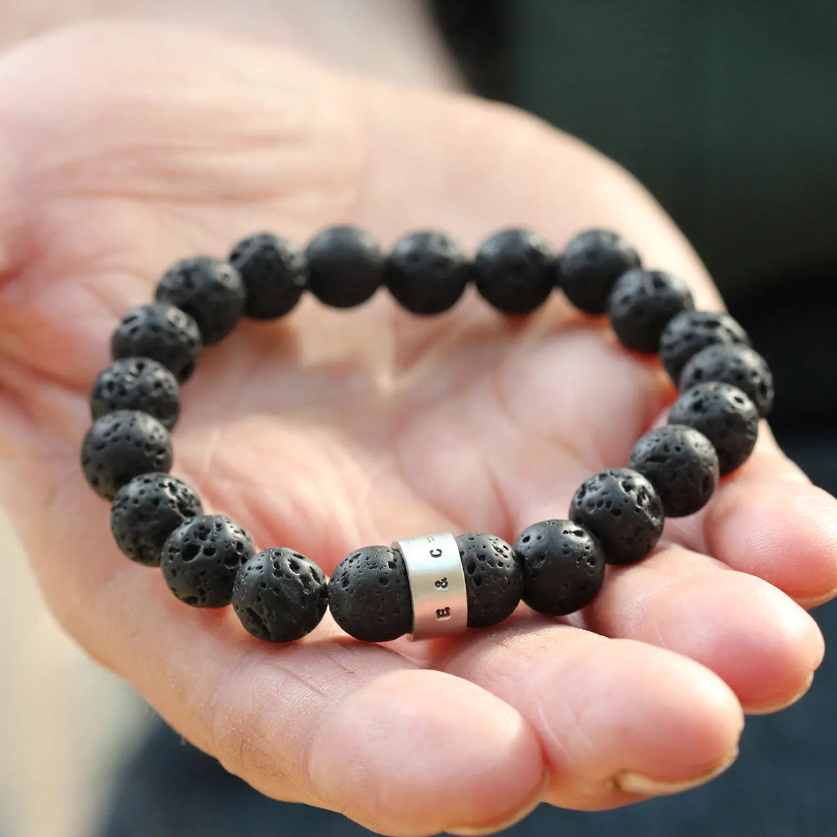 Close-up of black lava rock beads with natural porous surfaces and a satin silver hoop personalised with the initials E and C.