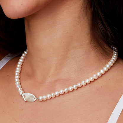 Model wearing a creamy pearl beaded necklace with a front T-Bar fastening and an oval disc which has the initial 'G' handstamped.