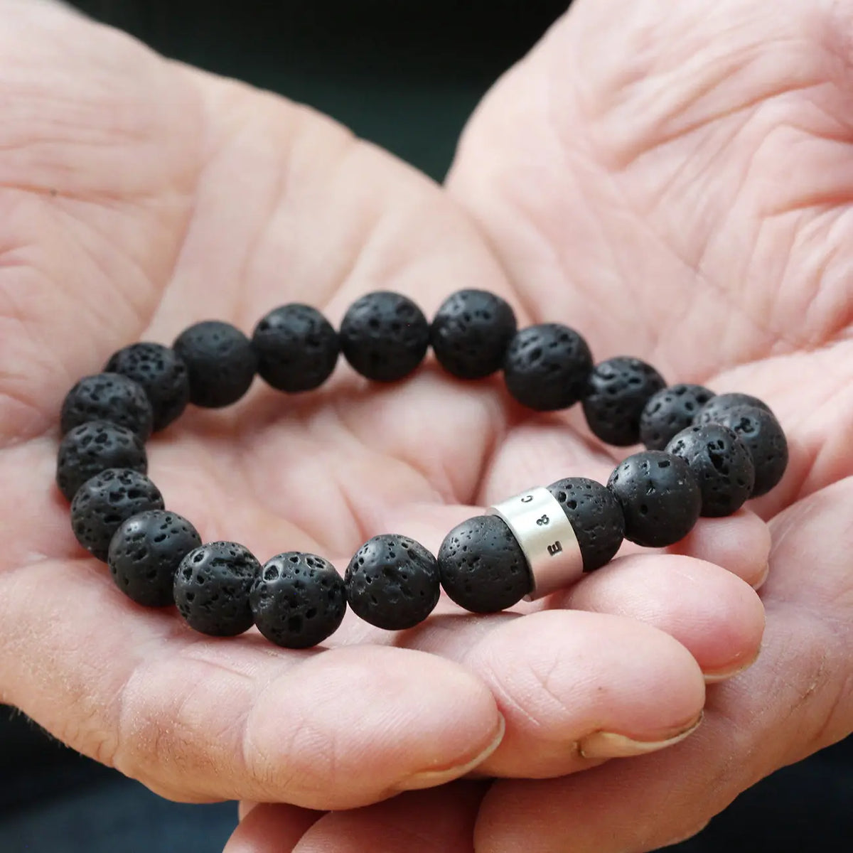 Personalised men's lava stone bracelet held in cupped hands, showing round matt black volcanic beads and a hand-punched silver hoop.