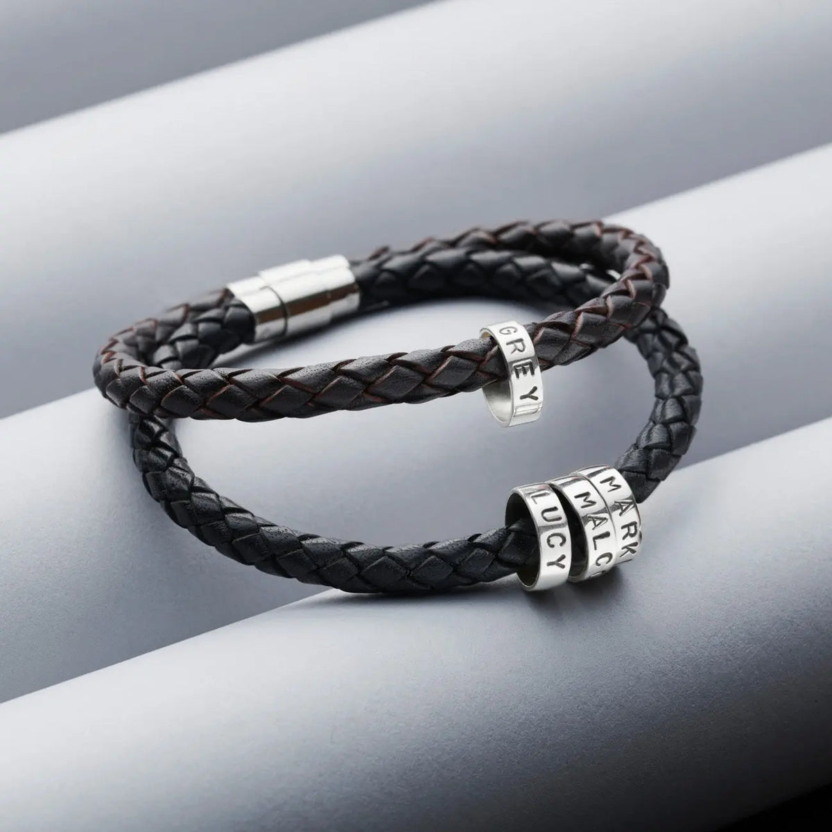 Black and dark brown men's braided leather bracelets with personalised sterling silver hoops and polished stainless steel clasps on a grey background.
